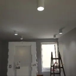 Zamontowane białe lampy sufitowe typu tuba w szarym pomieszczeniu w Żywcu, widoczna drabina i okno, drzwi zabezpieczone folią.