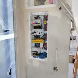 Wnęka w surowej ścianie z zamontowaną białą skrzynką rozdzielczą, zawierającą bezpieczniki i kolorowe kable. Nad skrzynką, w otworze okiennym widoczna izolacja z różową pianką. Po prawej stronie...