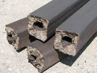 Stos brykietów Pini Kay o przekroju kwadratowym z charakterystycznym otworem w środku, leżących na szarym, betonowym podłożu.