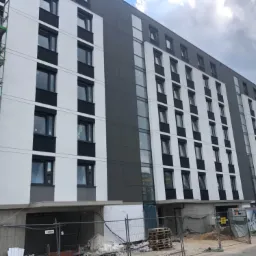 Wielopiętrowy budynek z częściowo ukończoną biało-szarą elewacją, widoczne rusztowania i teren budowy zabezpieczony siatką.