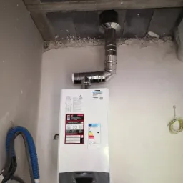 Nowy, biały gazowy kocioł kondensacyjny marki Ariston z podłączonym, chromowanym kominem, zamontowany na surowej ścianie z widocznymi przewodami instalacyjnymi.