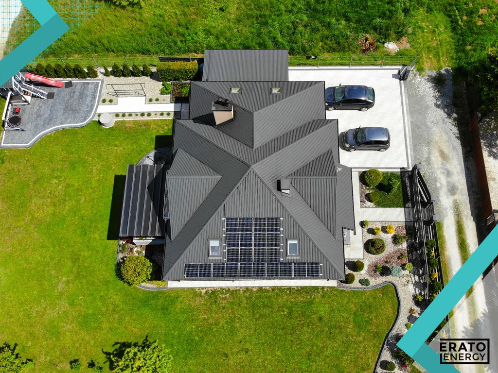 Widok z lotu ptaka na dom z szarym dachem i panelami słonecznymi, zadbany ogród z placem zabaw, dwa samochody na podjeździe z kostki brukowej.