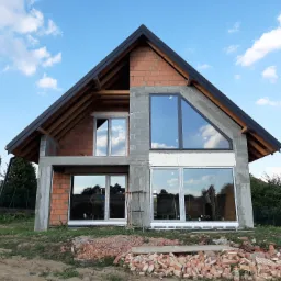 Surowy stan budynku jednorodzinnego z zamontowanymi oknami, widoczne bloczki betonowe, cegła dziurawka, biały styropian pod oknami, czarne orynnowanie i fragmenty rozbiórki cegieł na ziemi.