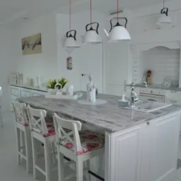 Jasna kuchnia w stylu rustykalnym z wyspą wykończoną blatem imitującym postarzane drewno, wiszące lampy w kształcie czajników i krzesła barowe z kwiatowym wzorem.