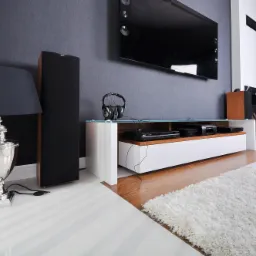 Nowoczesny salon z szarą ścianą, wiszącym telewizorem, białą szafką RTV z drewnianymi akcentami, kolumną głośnikową, srebrną lampą stołową i puszystym białym dywanem.