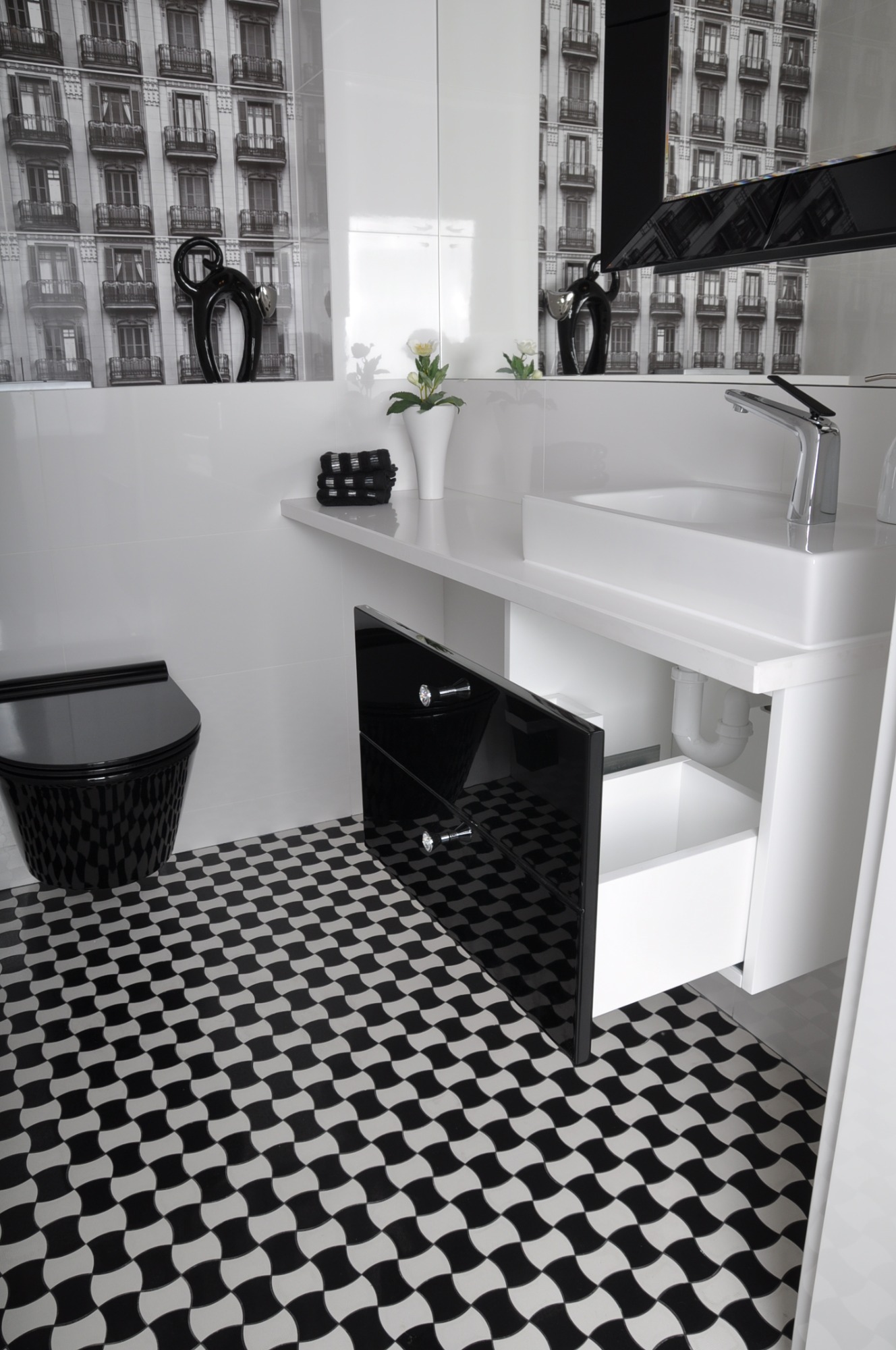 Nowoczesna łazienka w czarno-białej kolorystyce: biała umywalka nablatowa z chromowaną baterią, czarny sedes wiszący i otwarta szafka z czarnymi frontami na tle płytek z geometrycznym wzorem.