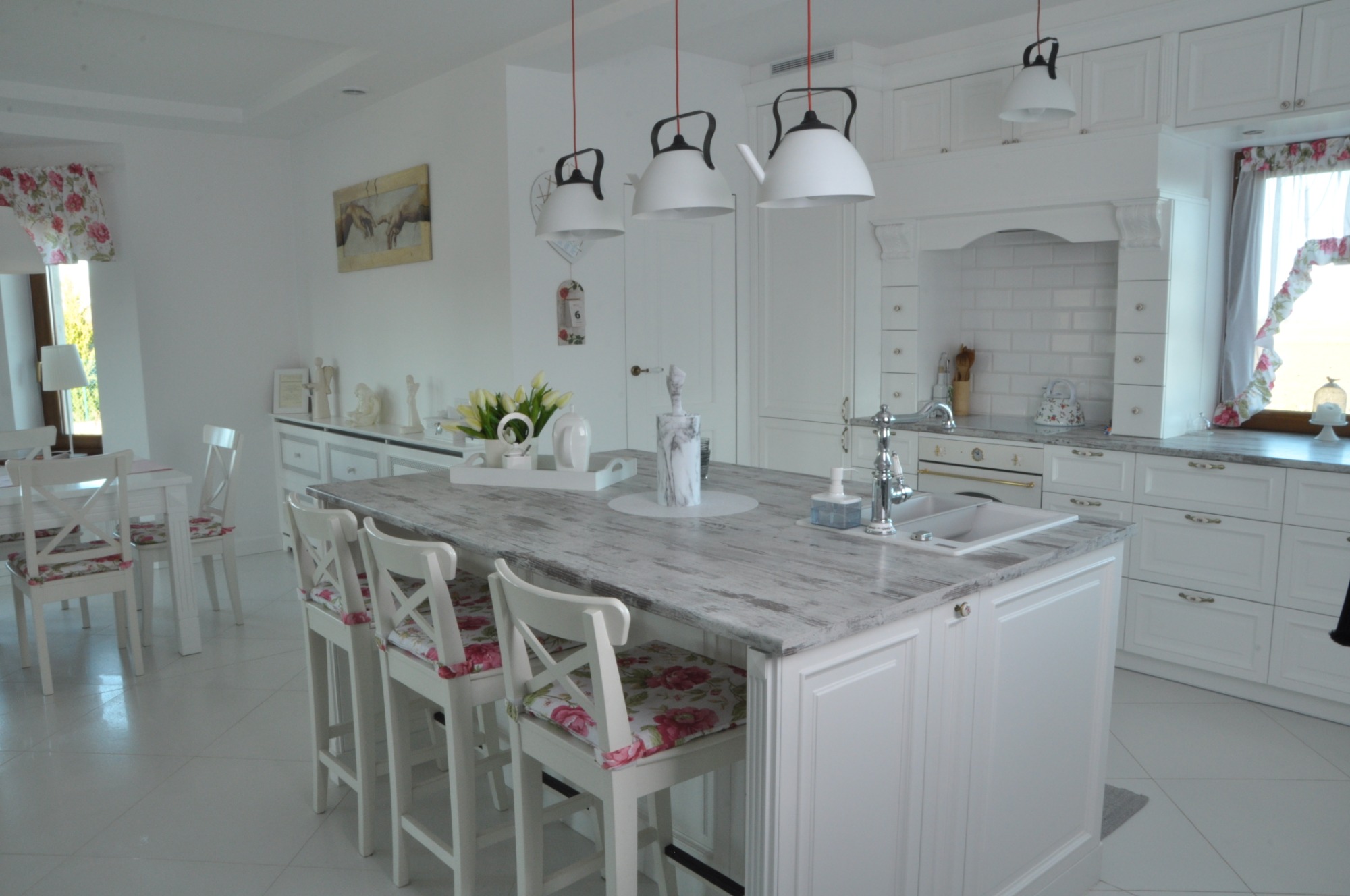 Jasna kuchnia w stylu rustykalnym z wyspą wykończoną blatem imitującym postarzane drewno, wiszące lampy w kształcie czajników i krzesła barowe z kwiatowym wzorem.