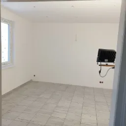 Pusty pokój po remoncie z nową, jasną podłogą wyłożoną płytkami imitującymi drewno, świeżo pomalowanymi białymi ścianami i widocznym oświetleniem roboczym.