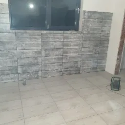 Ściana i podłoga wyłożone płytkami imitującymi drewno w szarym odcieniu, widoczna puszka elektryczna i zwinięty przedłużacz, okno z czarną ramą.