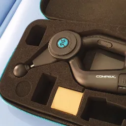 Czarny pistolet do masażu Compex w czarnym etui z pianki, widoczne różne nasadki, na niebieskim tle.