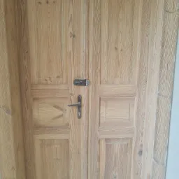 Drewniane dwuskrzydłowe drzwi po delikatnym oczyszczeniu, z widoczną strukturą drewna i oryginalnym zamkiem na środku.