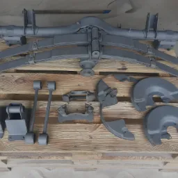 Szare elementy zawieszenia samochodowego, w tym osie, resory i osłony hamulcowe, ułożone na drewnianej palecie na piasku.