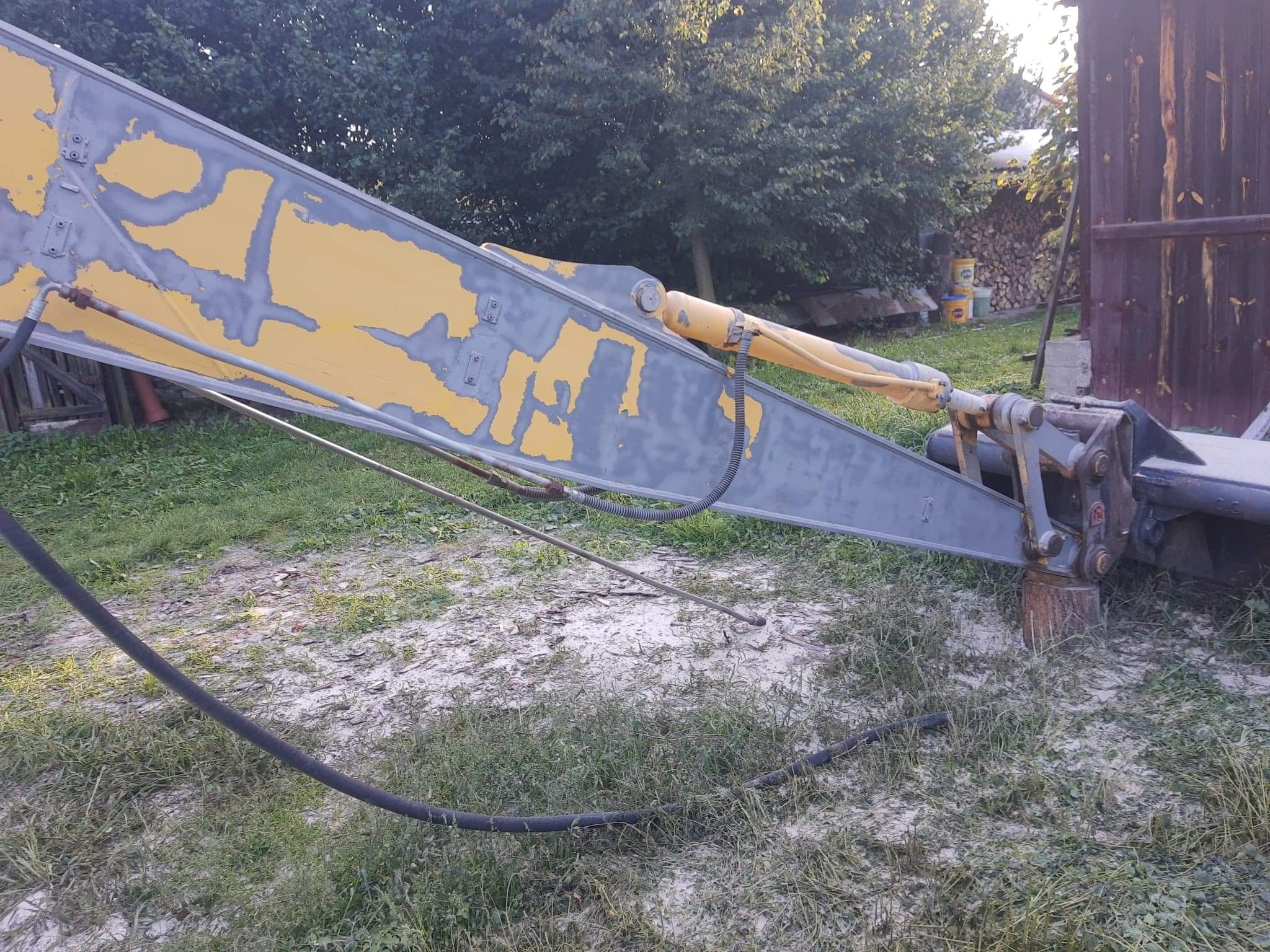 Ramię koparki z resztkami żółtej farby, przygotowane do ponownego malowania, widoczne elementy hydrauliczne i przewody.