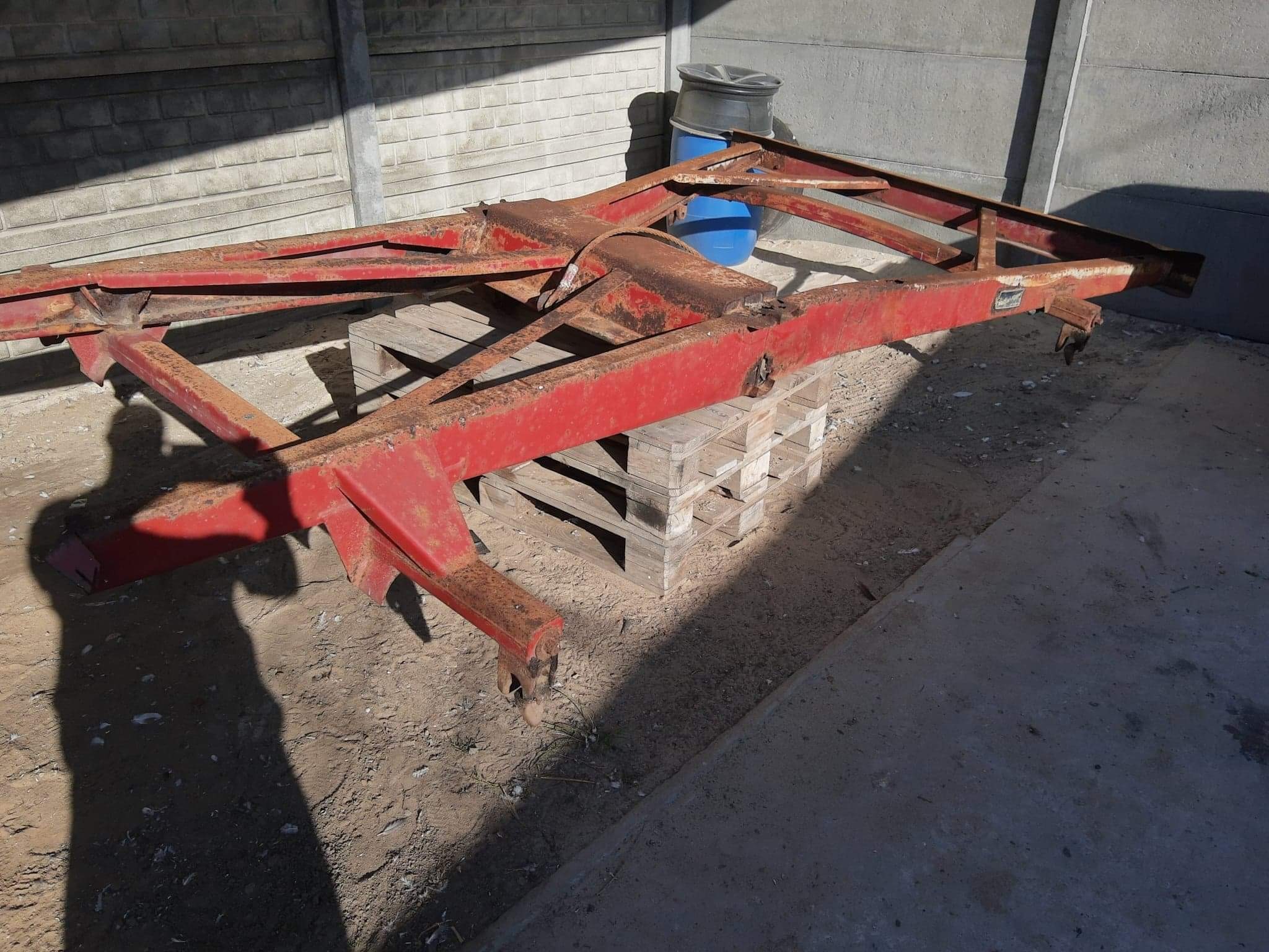 Częściowo skorodowana, czerwona, stalowa rama konstrukcyjna leżąca na palecie drewnianej, z widocznym cieniem osoby fotografującej i niebieskim pojemnikiem w tle.