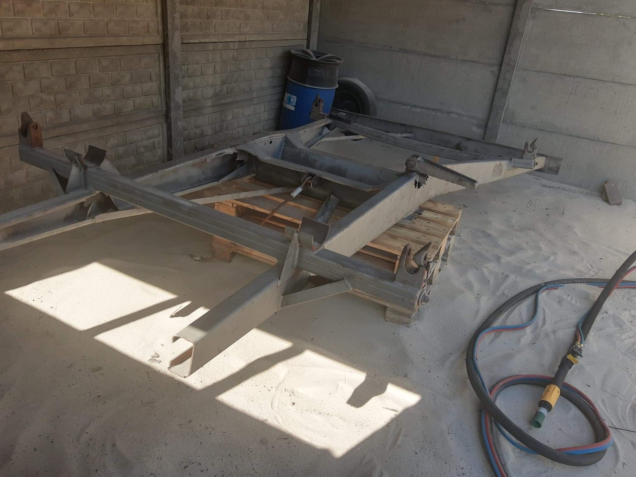 Metalowa rama przyczepy po obróbce, leżąca na drewnianej palecie, z widocznym wężem do piaskowania i betonowym tłem.