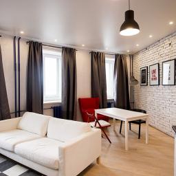 NORD-WEST Sp. z o. o. - Jasny salon z białymi ścianami, ceglaną ścianą akcentującą, drewnianą podłogą, szarymi zasłonami, białym narożnikiem i czarno-białym dywanem w szachownicę.