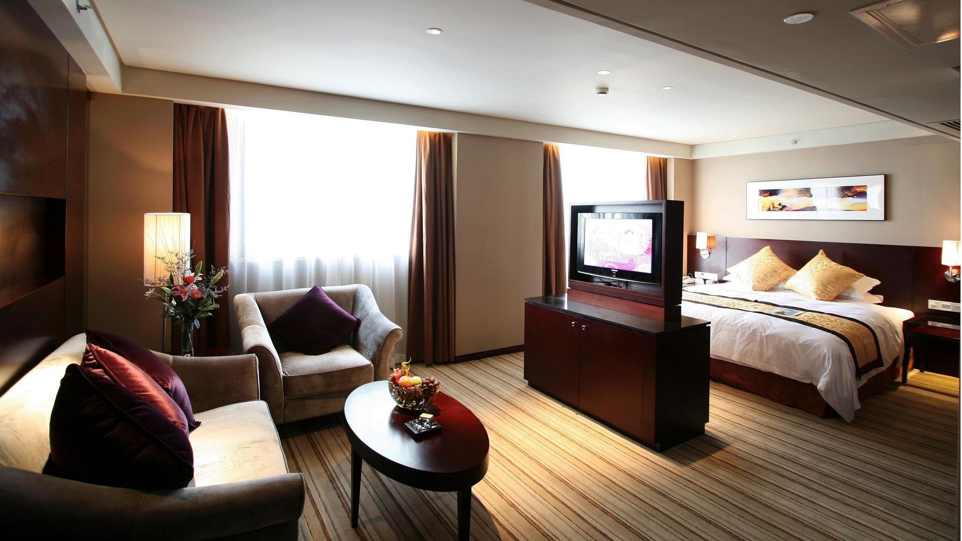 Elegancko urządzone wnętrze pokoju hotelowego z beżowymi ścianami, dwoma fotelami z fioletowymi poduszkami, owalnym stolikiem z owocami, telewizorem na komodzie oraz łóżkiem z żółtymi poduszkami...