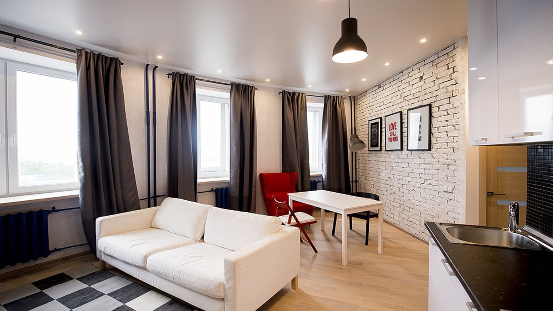Jasny salon z białymi ścianami, ceglaną ścianą akcentującą, drewnianą podłogą, szarymi zasłonami, białym narożnikiem i czarno-białym dywanem w szachownicę.