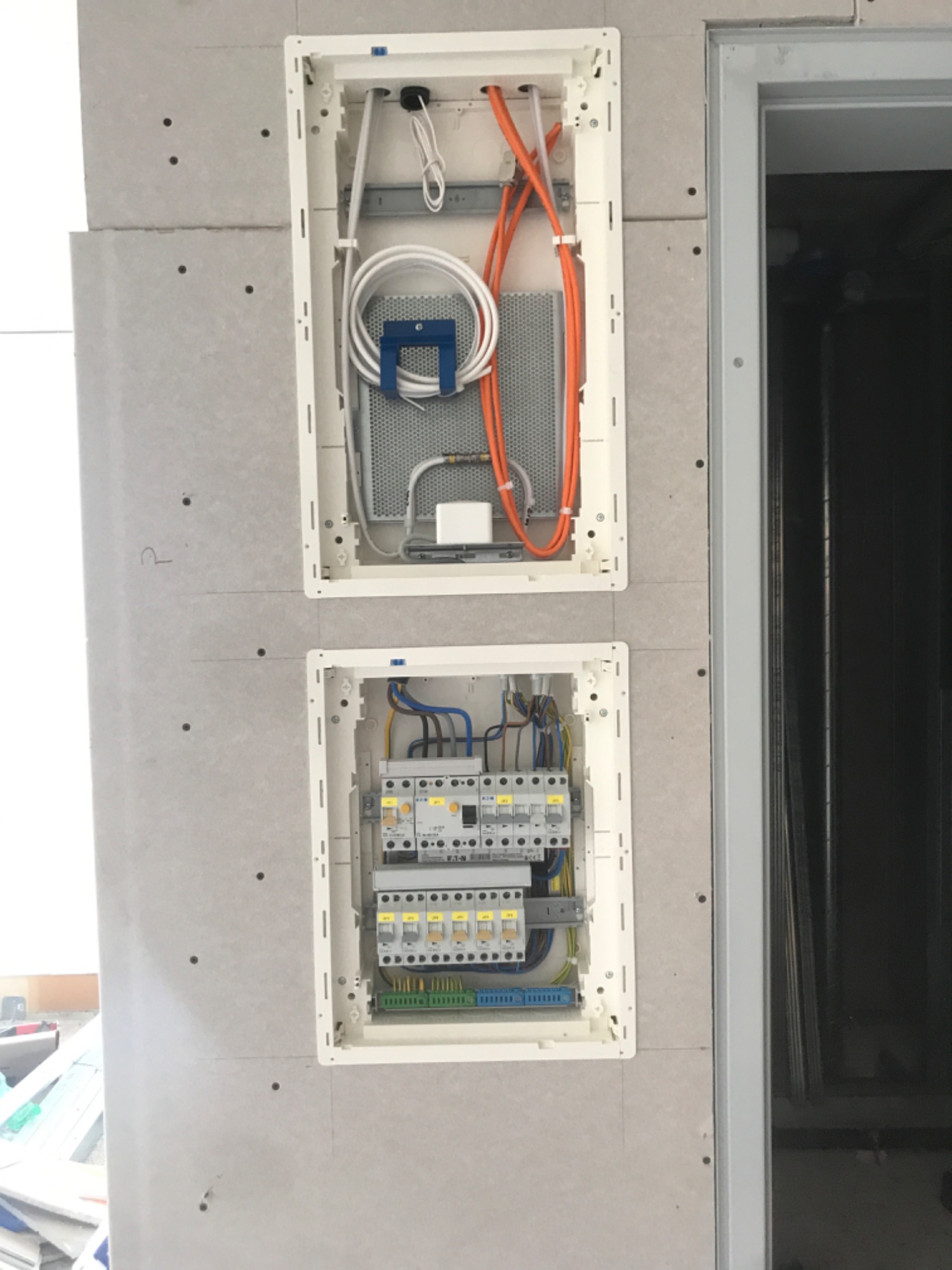 Dwa otwarte panele elektryczne w ścianie z płyt gipsowo-kartonowych w Nowym Sączu, górny z kablami telekomunikacyjnymi, dolny z bezpiecznikami i listwami zaciskowymi.