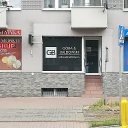 Fasada budynku z siedzibą kancelarii adwokackiej Góra & Baldowski, z logo GB na oknie i drzwiach, widoczne schody wejściowe i fragment przejścia dla pieszych. Na sąsiednim oknie widoczna reklama...
