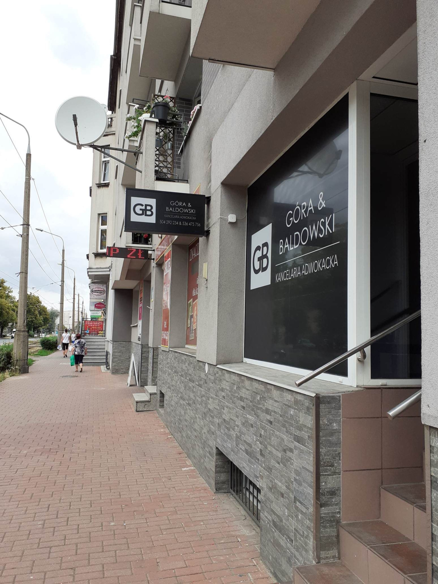 Fasada budynku z szyldem Kancelarii Adwokackiej Góra & Baldowski z widocznym logo GB na tle chodnika z kostki brukowej i przechodzących osób.