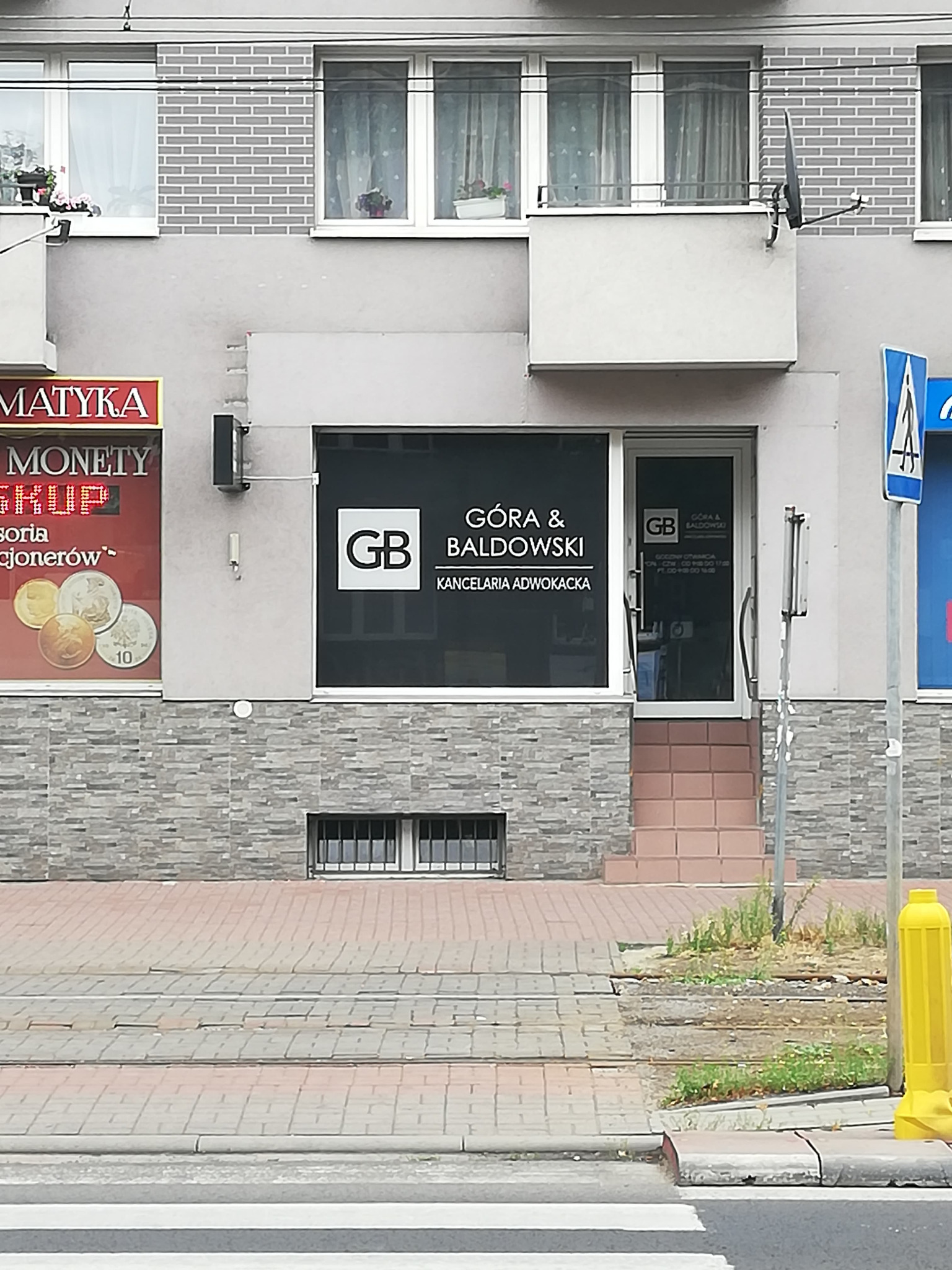 Fasada budynku z siedzibą kancelarii adwokackiej Góra & Baldowski, z logo GB na oknie i drzwiach, widoczne schody wejściowe i fragment przejścia dla pieszych. Na sąsiednim oknie widoczna reklama...