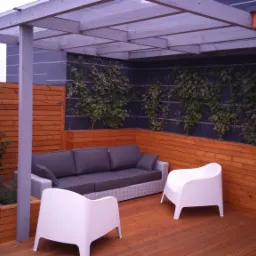 Zadaszony taras na dachu budynku z drewnianą podłogą i ścianami, wyposażony w szarą sofę z poduszkami, białe krzesła z tworzywa sztucznego oraz rośliny pnące się po ścianie.