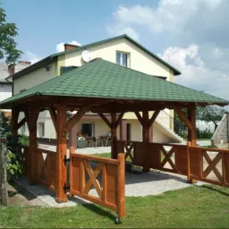 Drewniana altana z zielonym dachem z gontów bitumicznych, częściowo zabudowana niskimi płotkami z motywem krzyża, na tle domu jednorodzinnego z jasną elewacją i ogrodem.
