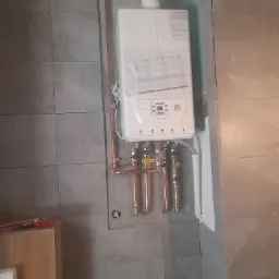 Biały piec gazowy Immergas zamontowany na szarej, kafelkowanej ścianie, z widocznymi miedzianymi rurami i zaworami, w trakcie instalacji, z niezakończonymi pracami wykończeniowymi wokół komina.