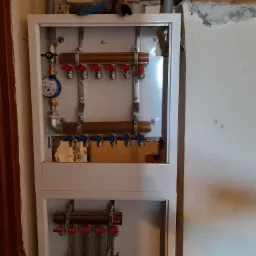 Rozdzielacz ogrzewania podłogowego w szafce, z widocznym licznikiem zużycia wody i zaworami regulacyjnymi, zamontowany na tle ściany z uszkodzonym tynkiem.