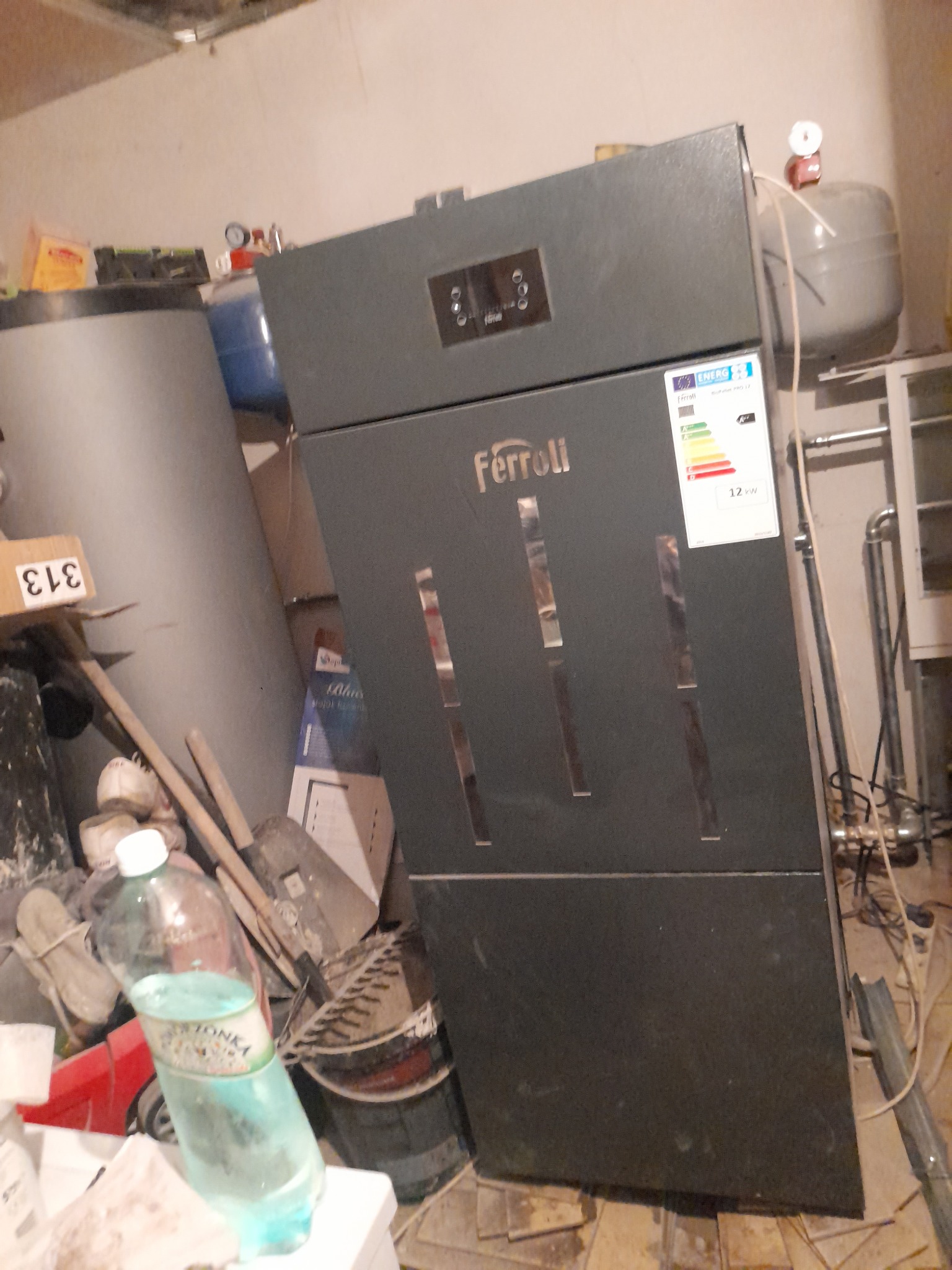 Szary piec grzewczy Ferroli o mocy 12kW w kotłowni, widoczna butelka wody mineralnej, rury i narzędzia w tle.