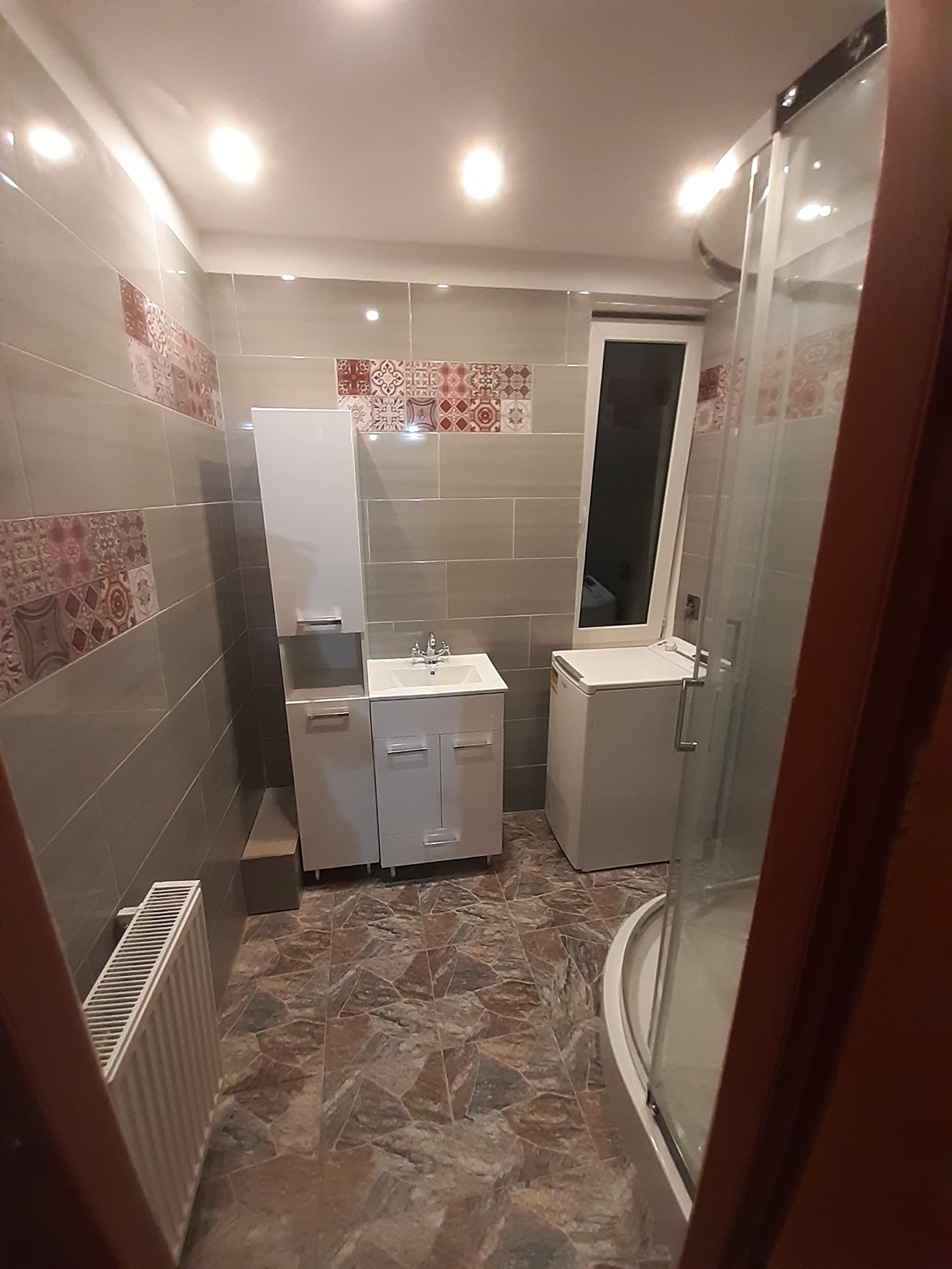 Ukończona łazienka z szarą glazurą, dekoracyjnymi bordiurami, białą zabudową umywalki i kabiną prysznicową z zaokrąglonym profilem, widoczny grzejnik żeberkowy.