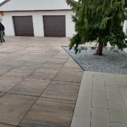 Ułożona kostka brukowa w odcieniach brązu i beżu przed garażem z brązowymi bramami, obok iglaste drzewo z szarymi kamieniami. Widoczne fragmenty osoby.