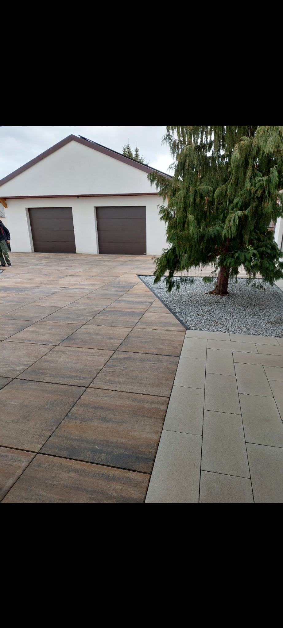 Ułożona kostka brukowa w odcieniach brązu i beżu przed garażem z brązowymi bramami, obok iglaste drzewo z szarymi kamieniami. Widoczne fragmenty osoby.