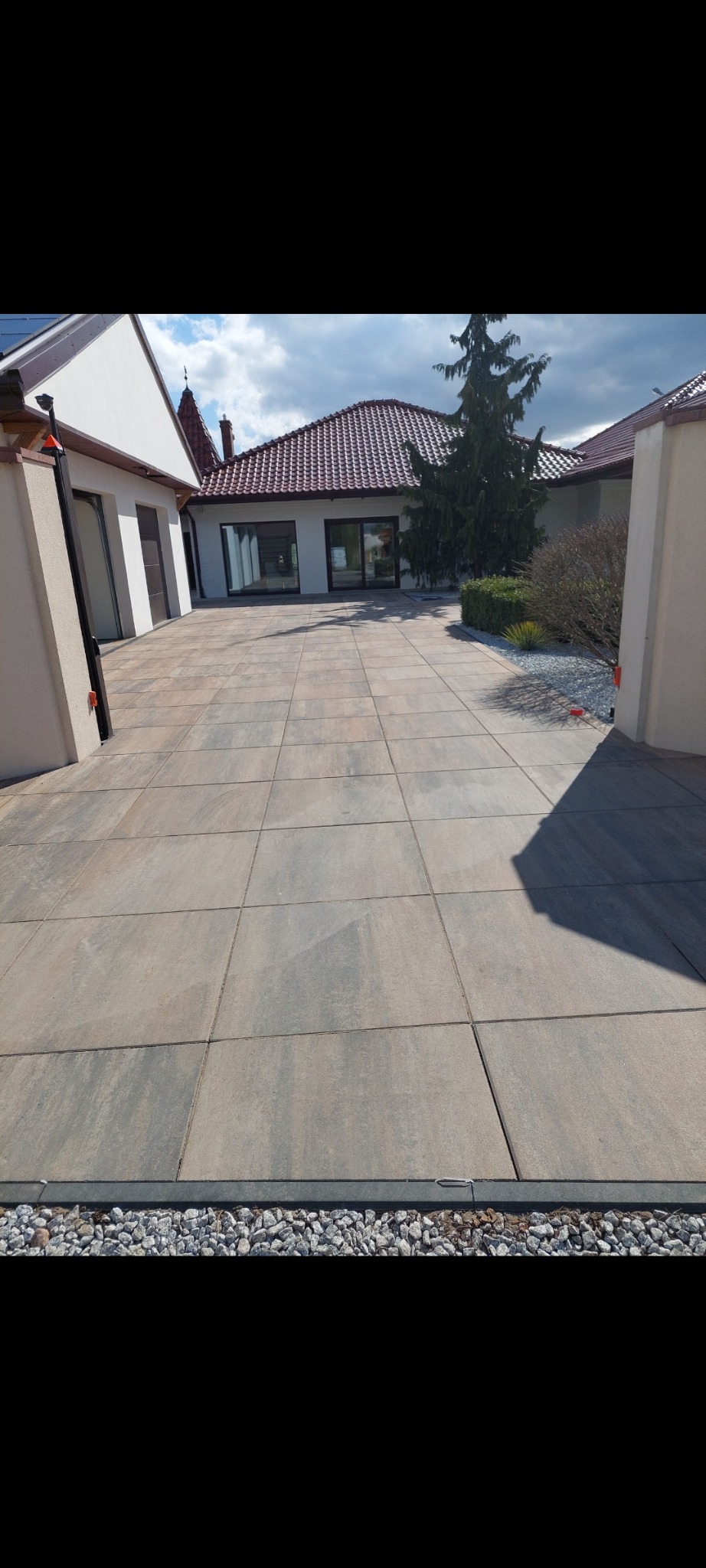 Ułożona kostka brukowa na podjeździe w Dziewierzewie, widok na dom z czerwonym dachem. Obrzeża z jasnych kamieni. Widoczne krzewy i drzewo iglaste.