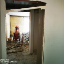 Wnętrze w trakcie tynkowania maszynowego: widoczny agregat tynkarski na kółkach, worki z zaprawą i świeżo nałożony tynk na ścianach w surowym stanie.