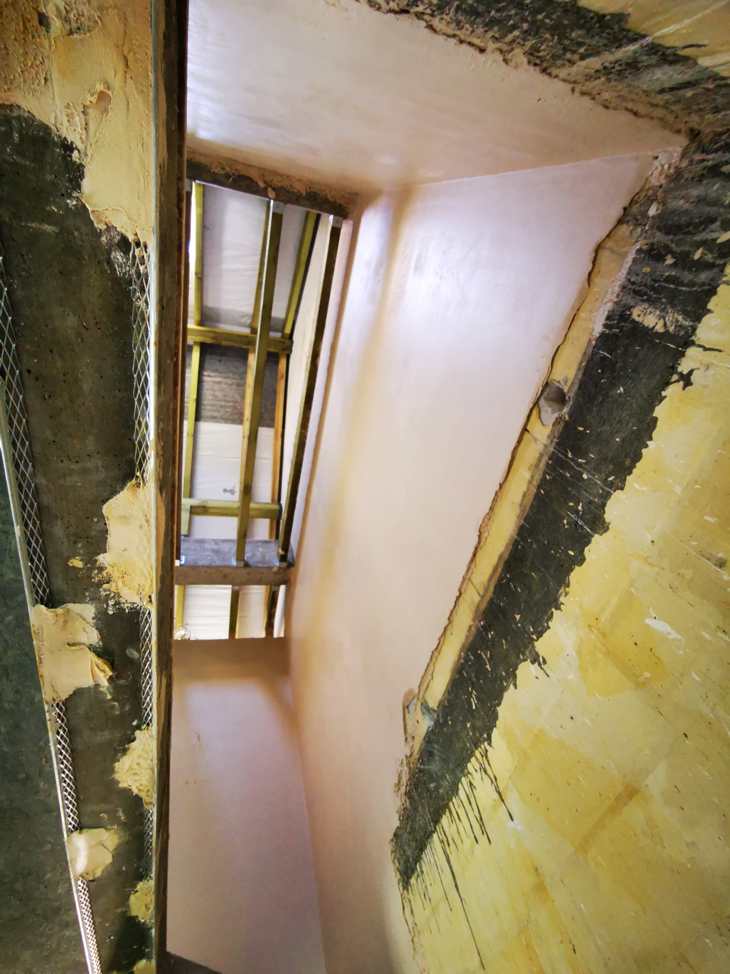 Wnętrze w trakcie prac wykończeniowych: ściana z nałożonym świeżym, jasnoróżowym tynkiem, obok fragment ściany z siatką i pozostałościami starego tynku, widoczne drewniane elementy konstrukcyjne...