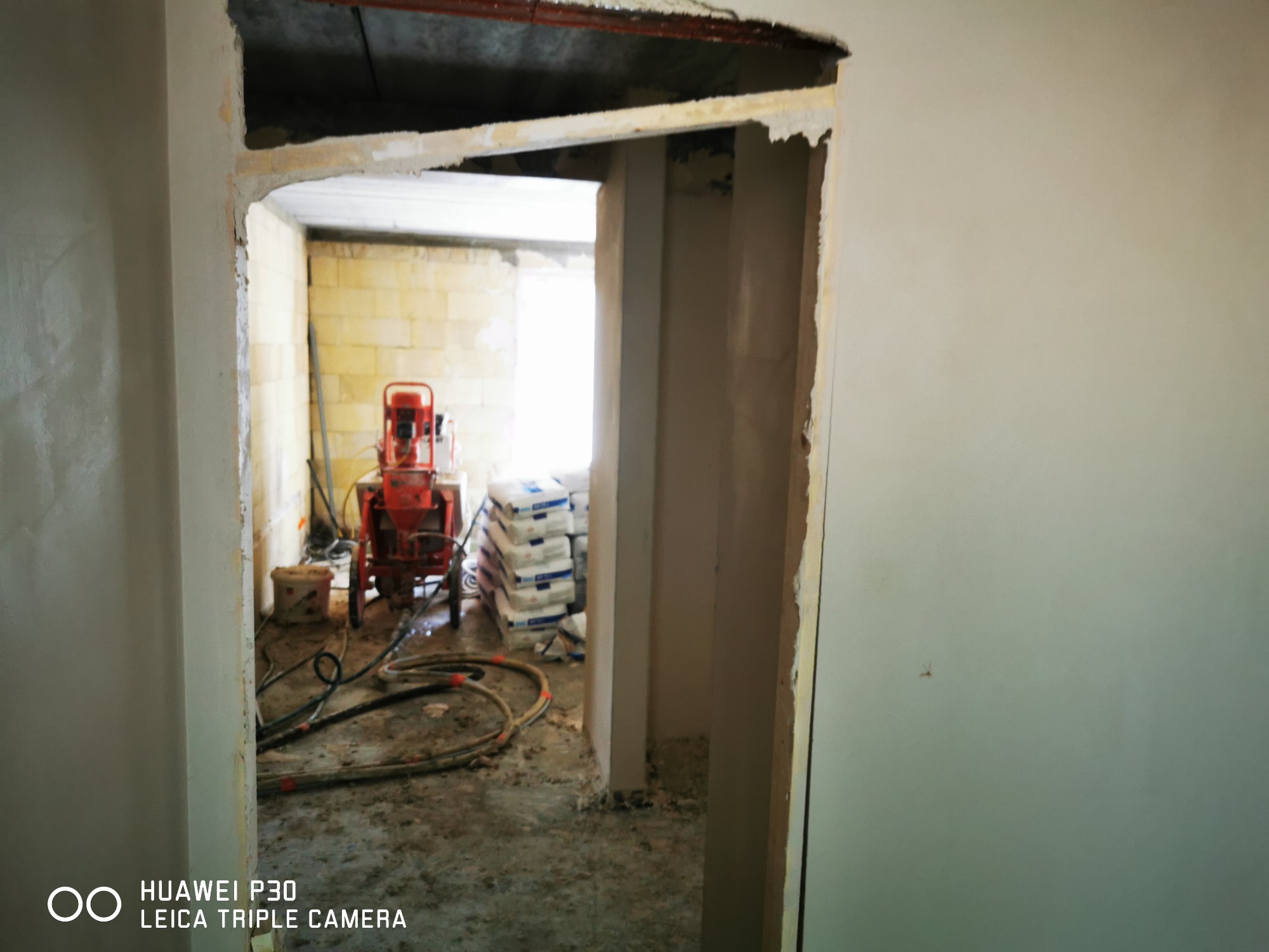 Wnętrze w trakcie tynkowania maszynowego: widoczny agregat tynkarski na kółkach, worki z zaprawą i świeżo nałożony tynk na ścianach w surowym stanie.