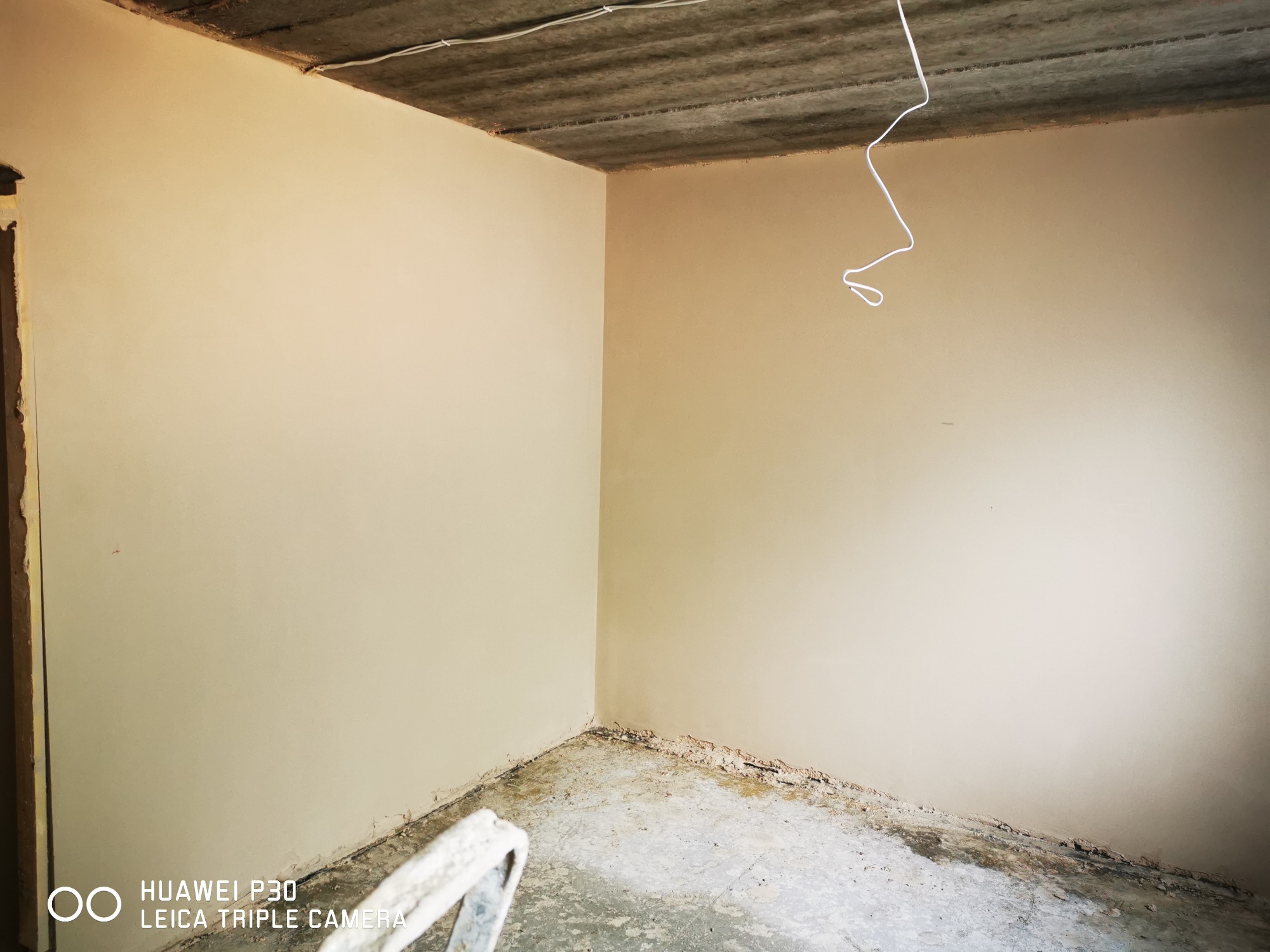 Wnętrze pomieszczenia po nałożeniu gładzi, widoczne świeżo otynkowane ściany w beżowym odcieniu i surowy betonowy sufit z wystającym kablem elektrycznym, w rogu fragment drabiny.