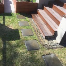 Ogrodowe Kurtyzany - Stopnie tarasowe z drewna prowadzące do podwyższonej rabaty, połączone ścieżką z kwadratowych płyt chodnikowych na trawniku.