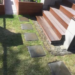 Stopnie tarasowe z drewna prowadzące do podwyższonej rabaty, połączone ścieżką z kwadratowych płyt chodnikowych na trawniku.