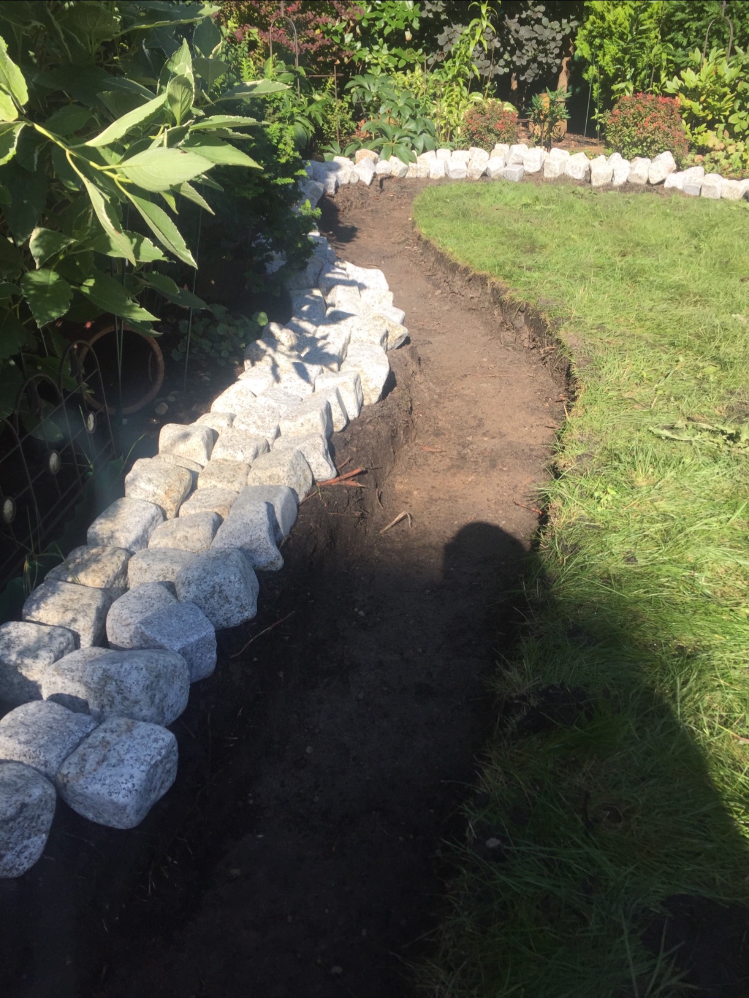 Krawężnik z jasnoszarych kamieni oddziela ścieżkę z ciemnej ziemi od trawnika w ogrodzie.