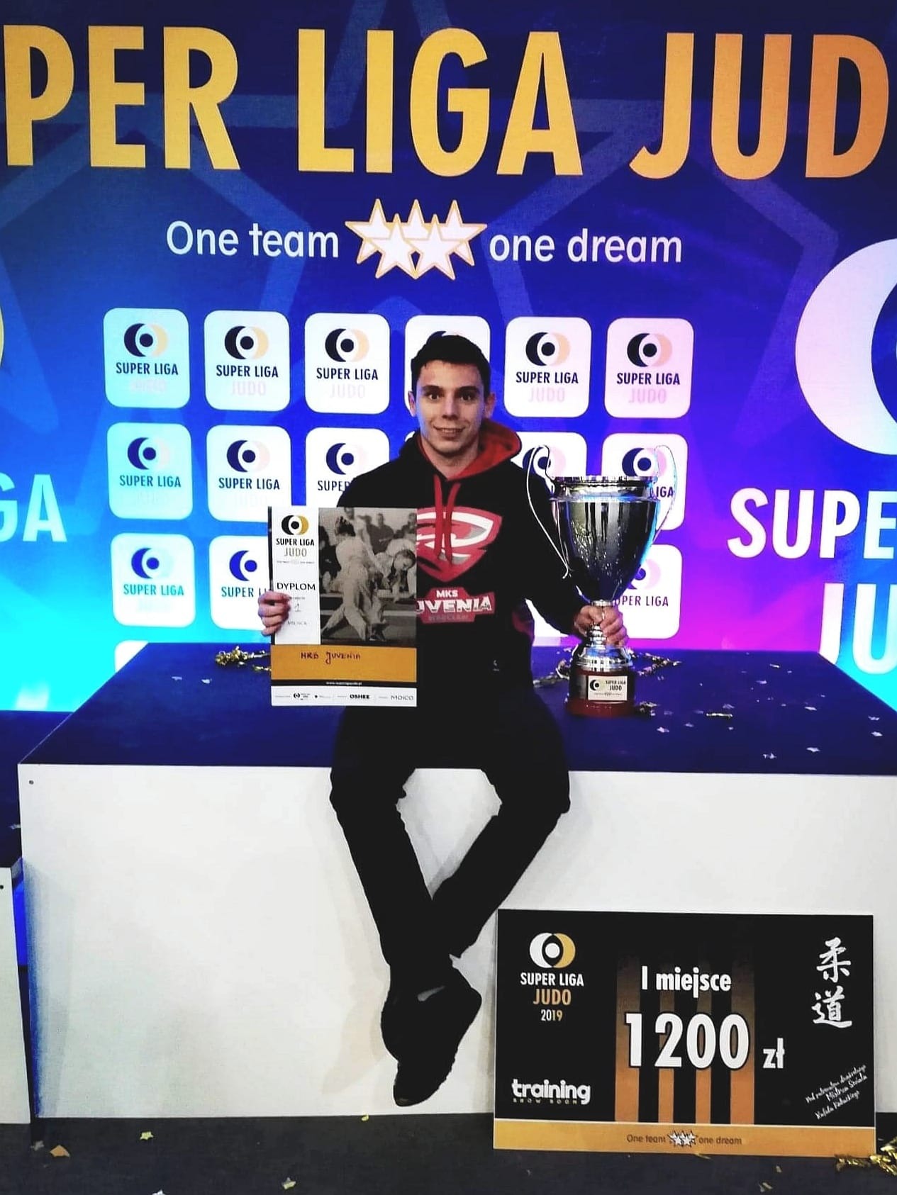 Judoka z pucharem i dyplomem za pierwsze miejsce w Super Lidze Judo, na tle baneru reklamowego z logo ligi i hasłem 'One team, one dream'.