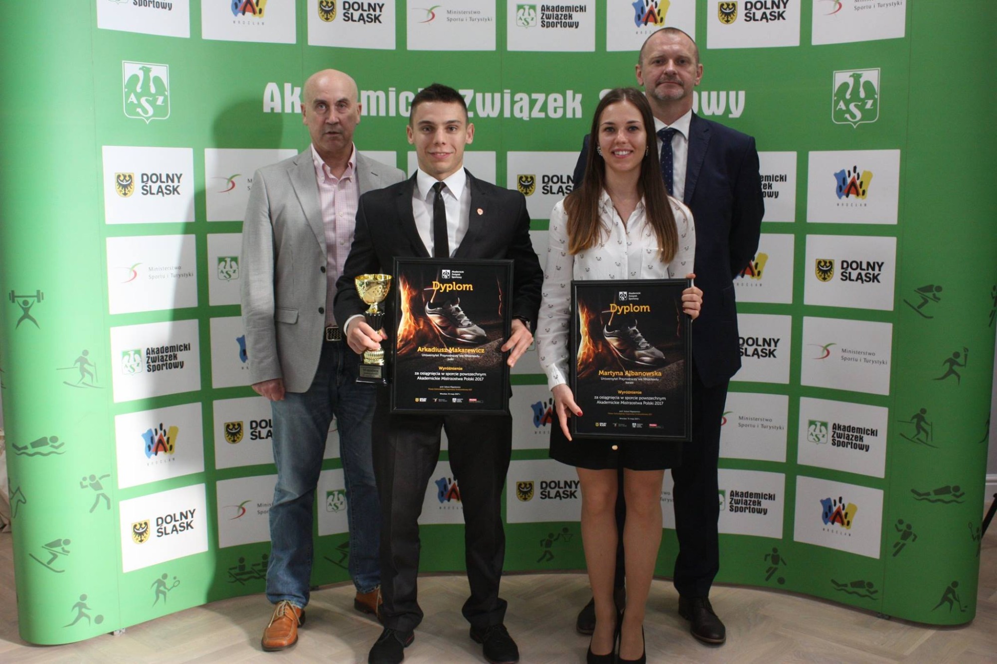 Grupa osób prezentuje dyplomy i puchar za osiągnięcia sportowe na tle ścianki z logotypami Akademickiego Związku Sportowego i Dolnego Śląska.