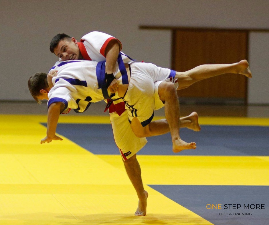 Dwóch mężczyzn w białych strojach z kolorowymi pasami ćwiczy rzut podczas treningu sportów walki na żółto-szarej macie.