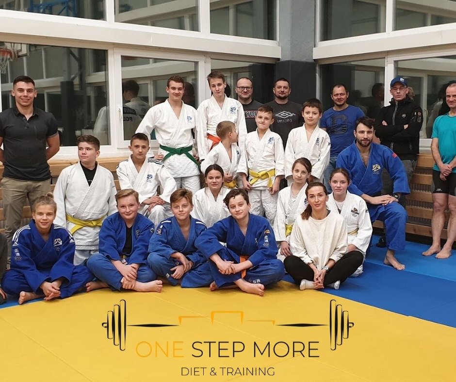 Grupa dzieci i dorosłych w strojach do judo, z różnymi kolorami pasów, pozuje na żółto-niebieskiej macie w sali treningowej, z logo 'One Step More Diet & Training' w dolnej części kadru.