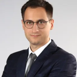 Portret młodego mężczyzny w okularach i granatowym garniturze w kratę, z szarym krawatem, na jasnoszarym tle.