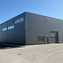 Mix-Bud - Nowoczesny, szary budynek przemysłowy z płyt warstwowych pod błękitnym niebem. Widoczne logo 'ENSTO' i dwie bramy wjazdowe.