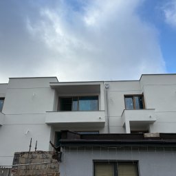 Mix-Bud - Nowoczesny, biały dom z balkonami i dużymi oknami, widoczny spod niższego budynku z elementami rozbiórki. Pochmurne niebo.