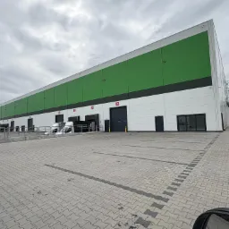 Nowoczesny budynek magazynowy z zielono-białą elewacją, widok z zewnątrz, z rampami załadunkowymi i dwoma białymi samochodami dostawczymi na brukowanym placu.